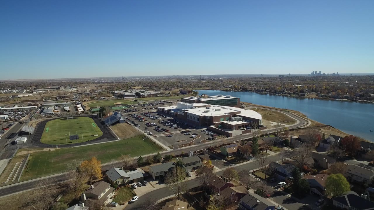 Aerial frame in Hidden Lake, Colorado, with the green field and parking area centered, the lake running along the right side, distant buildings on the horizon, and no visible people.