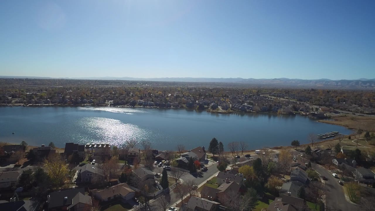 Wide aerial view of a lake, rooftops, streets, and distant hills under clear sky.