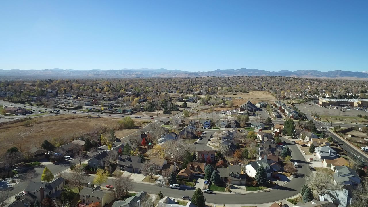 Aerial view of suburban homes, roads, fields, and distant mountains under clear sky.