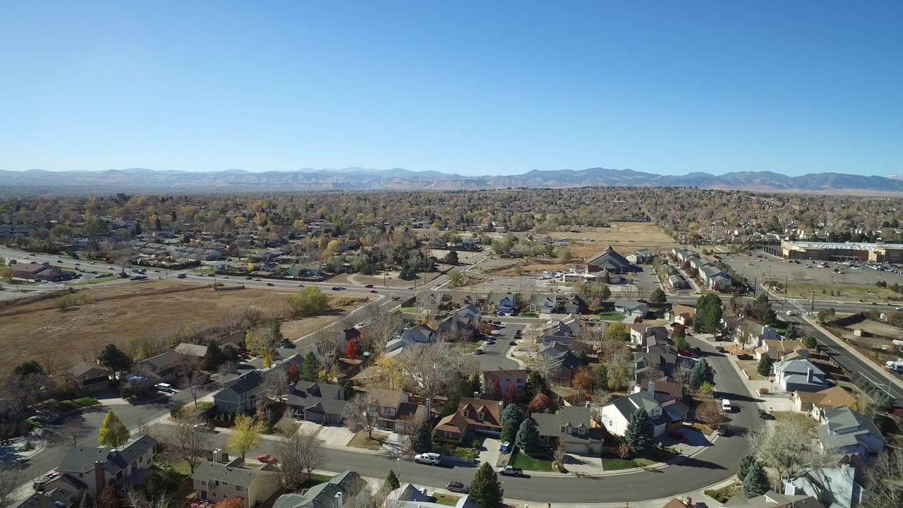 Aerial view in Hidden Lake, Colorado with houses in the foreground, a larger road and parking areas beyond, open lots, mountain ridges, and no visible people.