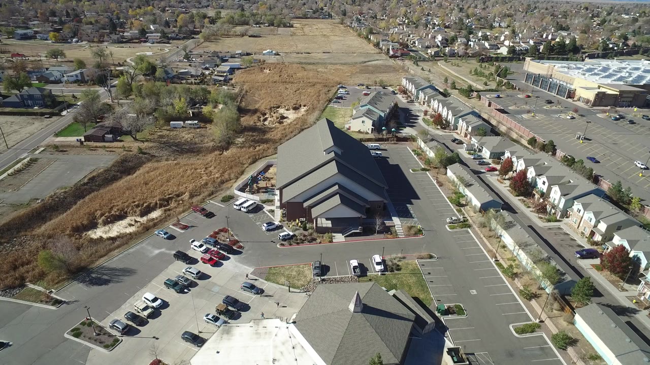 High aerial frame over Hidden Lake, Colorado, showing a dark-roofed building, parking areas with cars, dry open land, and a neighborhood in the distance.