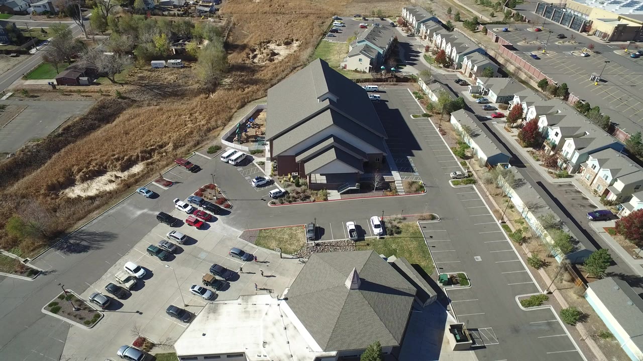 High aerial frame over Hidden Lake, Colorado showing a mostly full paved parking lot, a large dark-roofed building, townhomes along a road, dry open ground, and no visible people.