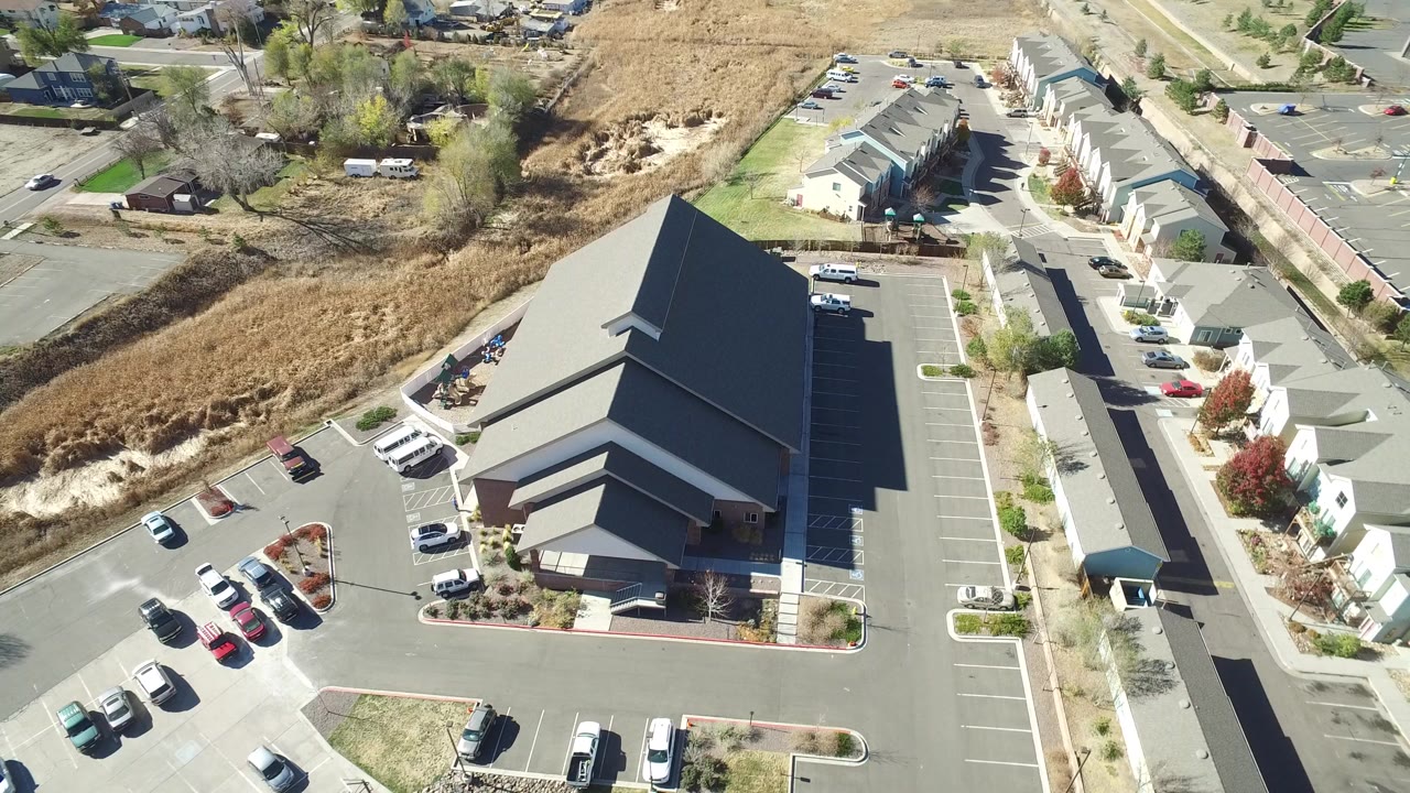 A high overhead frame in Hidden Lake, Colorado shows a large dark-roofed building, paved parking areas, parked cars, nearby houses, streets, dry grass, and no visible people.