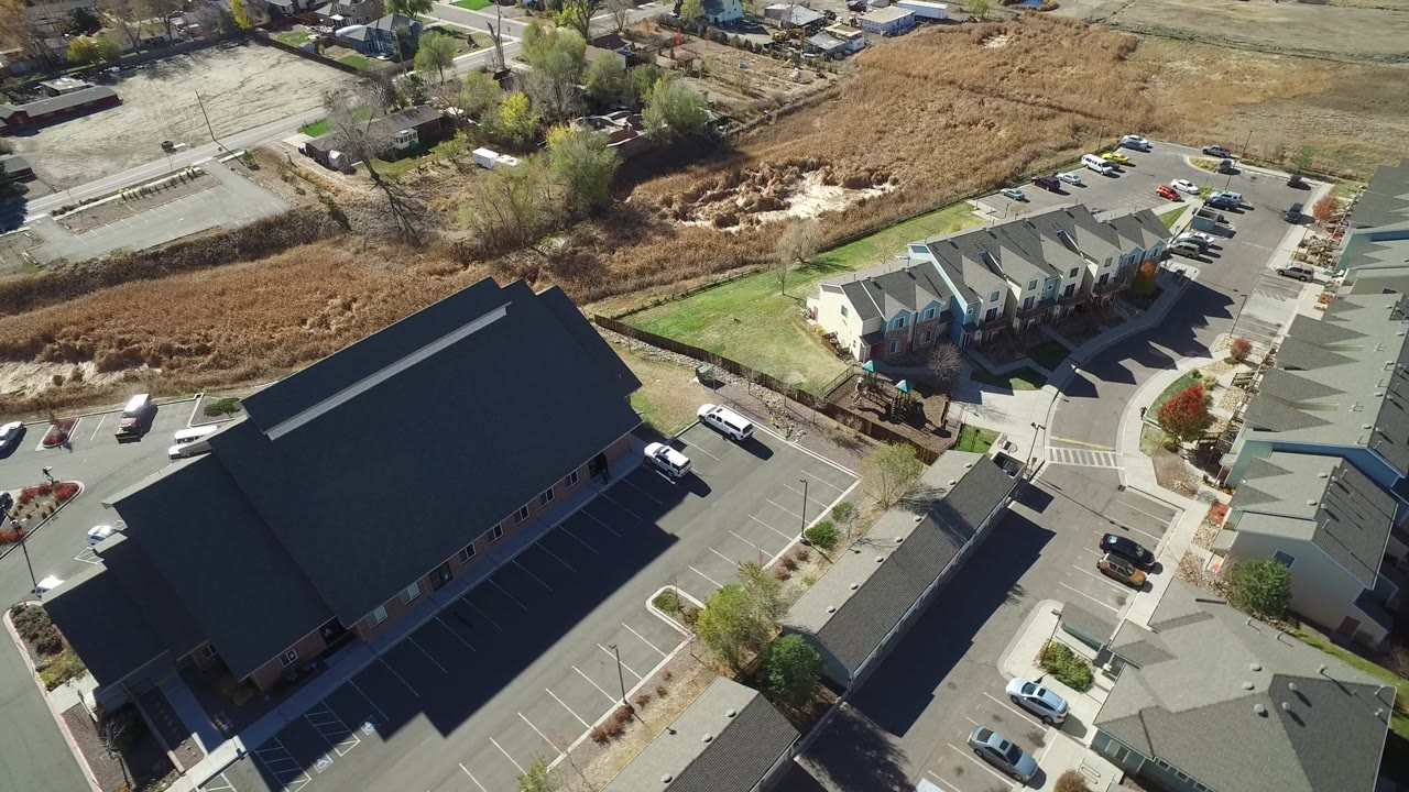 Aerial frame over Hidden Lake, Colorado with the dark-roofed building filling the left side, townhouses across the upper right, parked cars, crosswalk markings, and no visible people.