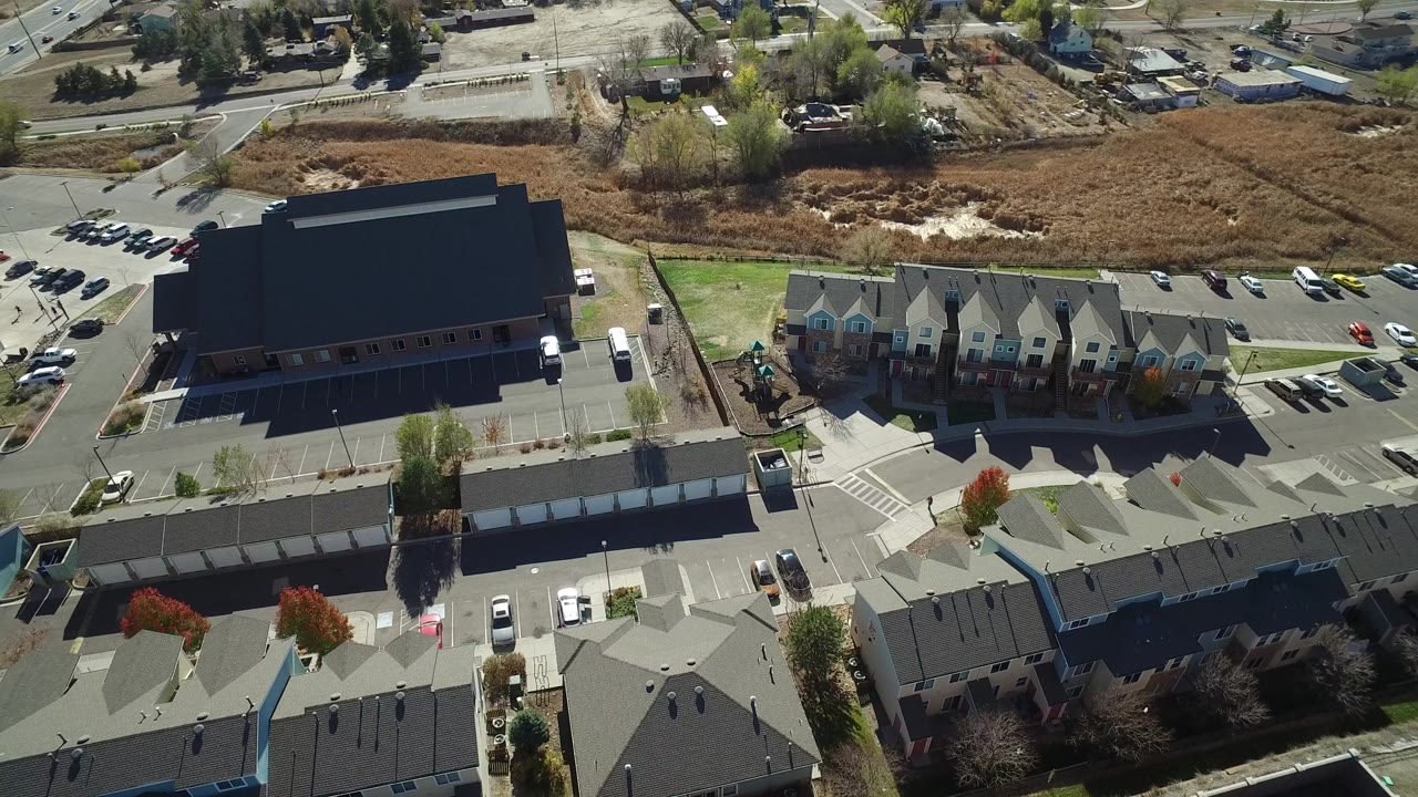 High aerial frame over Hidden Lake, Colorado showing a large dark-roofed building at left, townhome roofs at bottom and right, parking areas, roads, trees, and a brown field with no visible people.