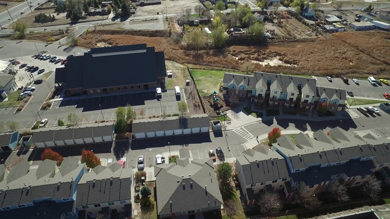 Overhead view of a black-roofed building, adjacent homes, parking areas, and roadside field.
