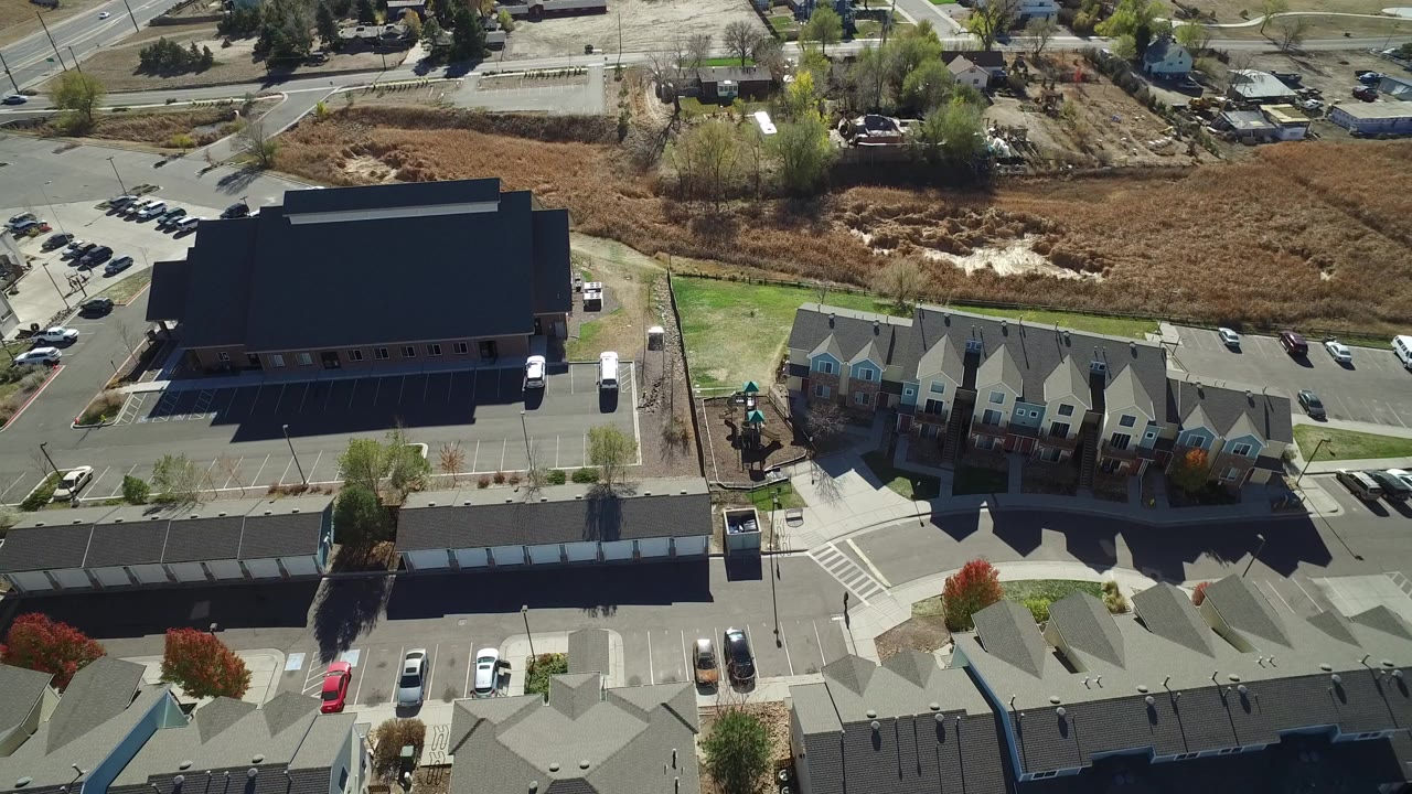 Aerial view of townhomes, a large dark-roofed building, parking lots, roads, and open brown land.
