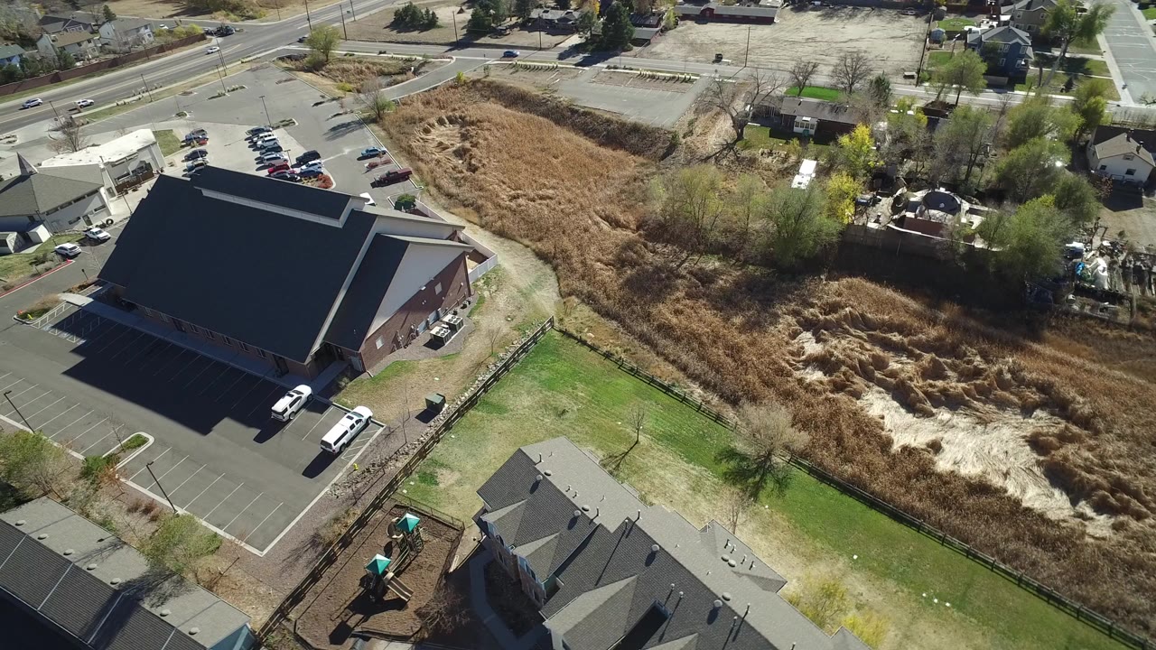 High aerial frame over Hidden Lake, Colorado showing a large dark-roofed building beside a parking lot, nearby roads, residential rooftops, fenced yards, tan scrubland, and no visible people.