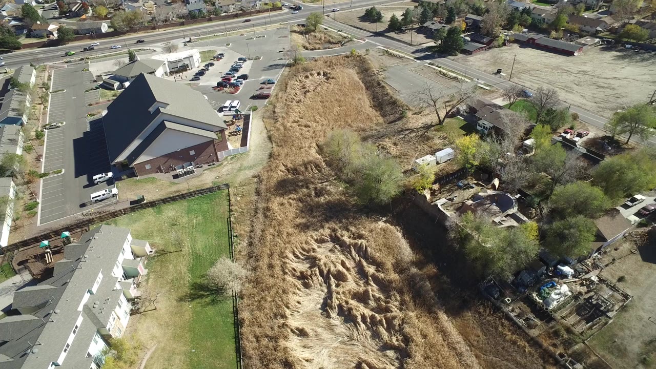 Overhead frame from Hidden Lake, Colorado showing white townhomes beside a green lawn, a large dark-roofed building, arterial roads, parking lots, and dry open parcels, with no visible people.