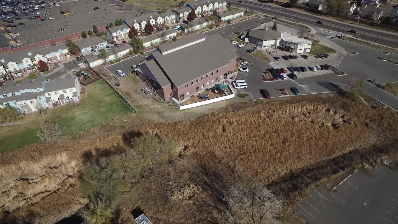 A high oblique drone frame in Hidden Lake, Colorado shows a large brick-toned building, a nearly full parking lot, tan marsh grass in the foreground, townhomes beyond the road, and no visible people.