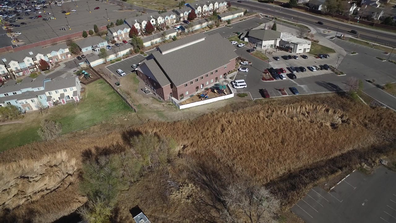 The Hidden Lake, Colorado aerial remains angled over the same large building and parking lot, with a strip of townhomes and a road across the upper frame and no visible people.