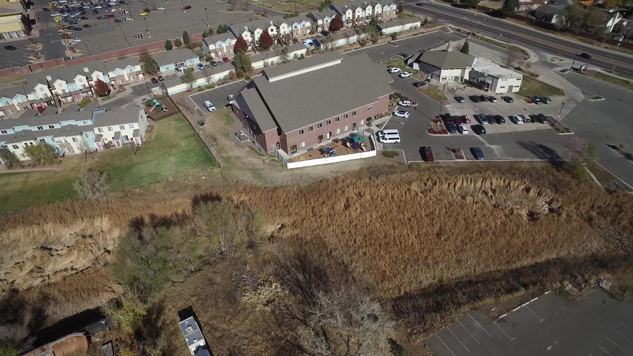 Aerial frame with parking lot, large roof, townhomes, road, and tan reeds.