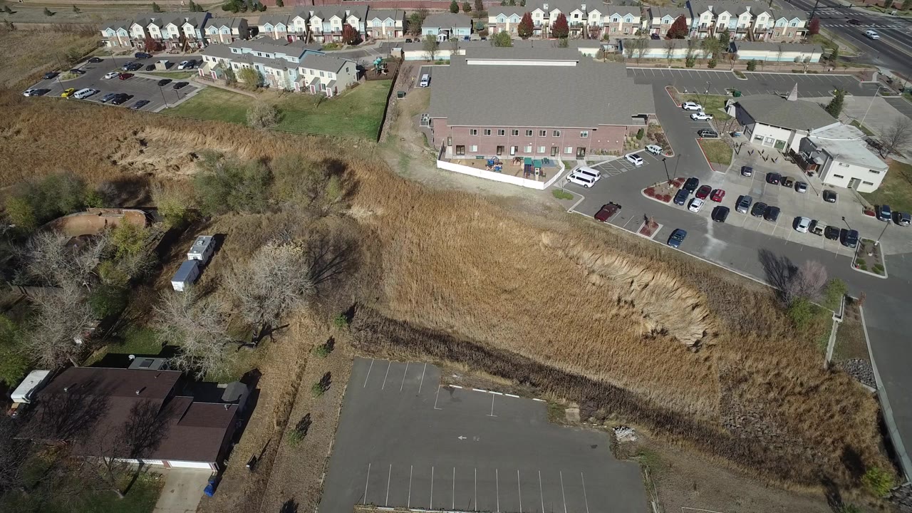 Aerial view in Hidden Lake, Colorado with the tan grass area centered, a paved lot at lower right, low buildings beyond it, homes along the top edge, and no visible people.