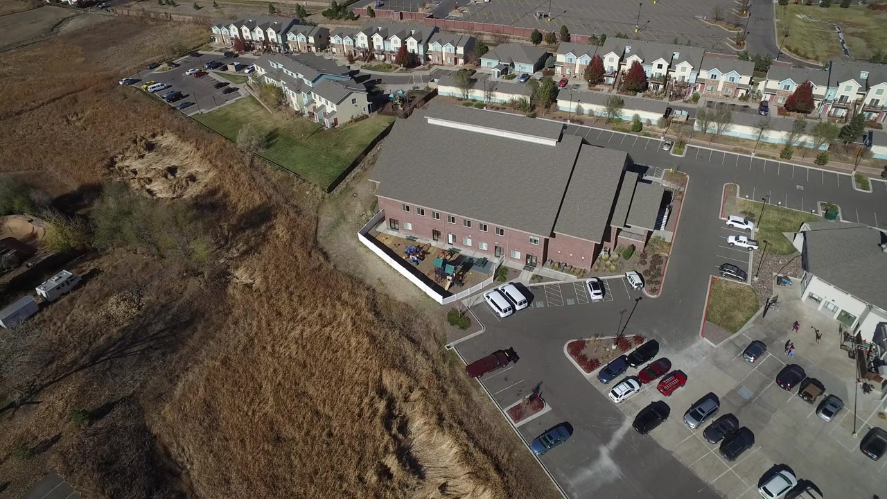 Aerial view of a large building, parking lot, homes, and dry open land.