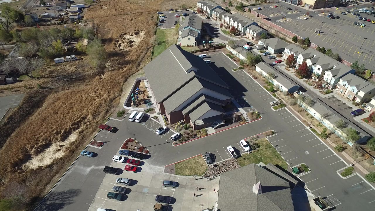 Opening aerial frame over a public chapel in Hidden Lake, Colorado, with paved lots, parked cars, townhomes beyond the road, dry open land at left, and no visible people.