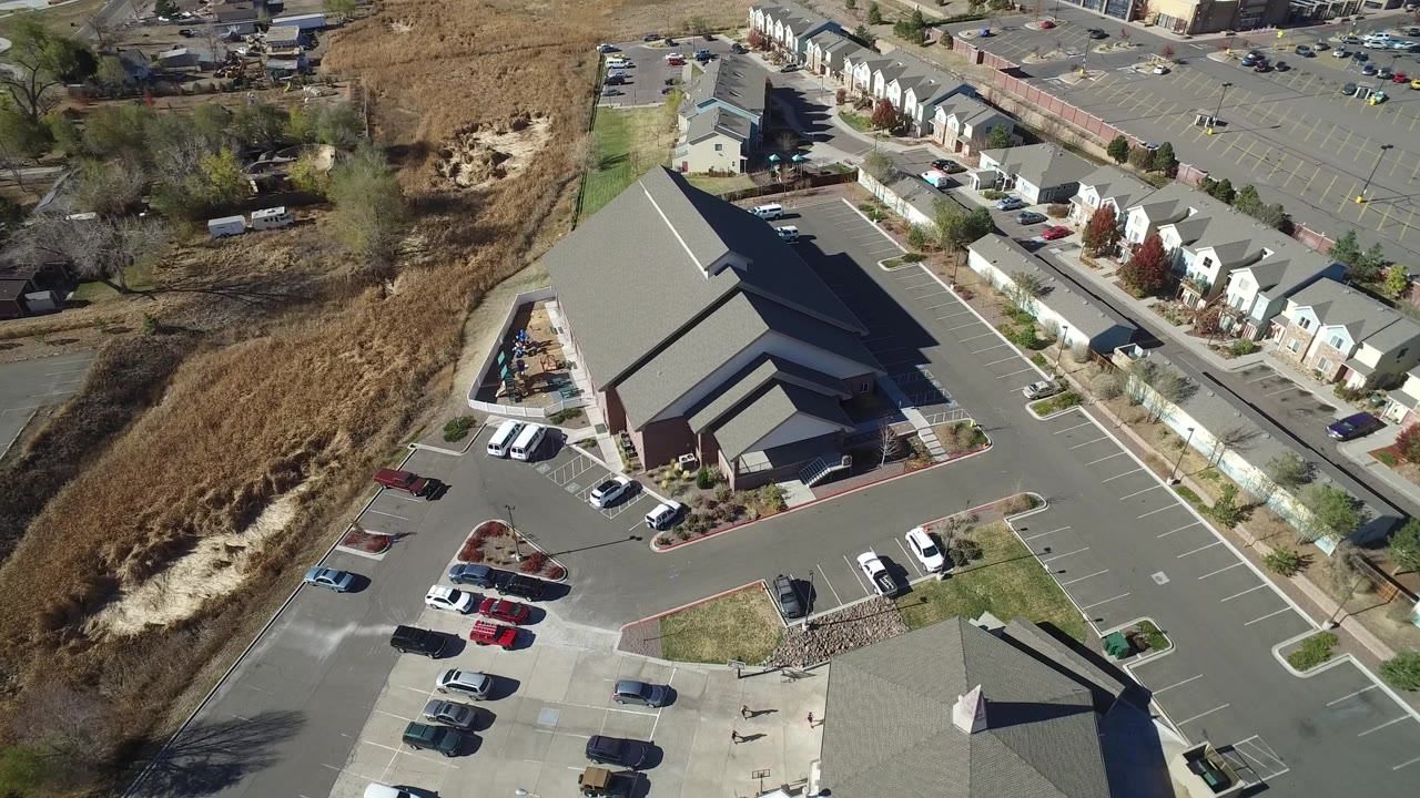 Aerial frame in Hidden Lake, Colorado showing the chapel roof and surrounding parking lots, with a row of townhomes along the road and dry grassland on the left, no visible people.