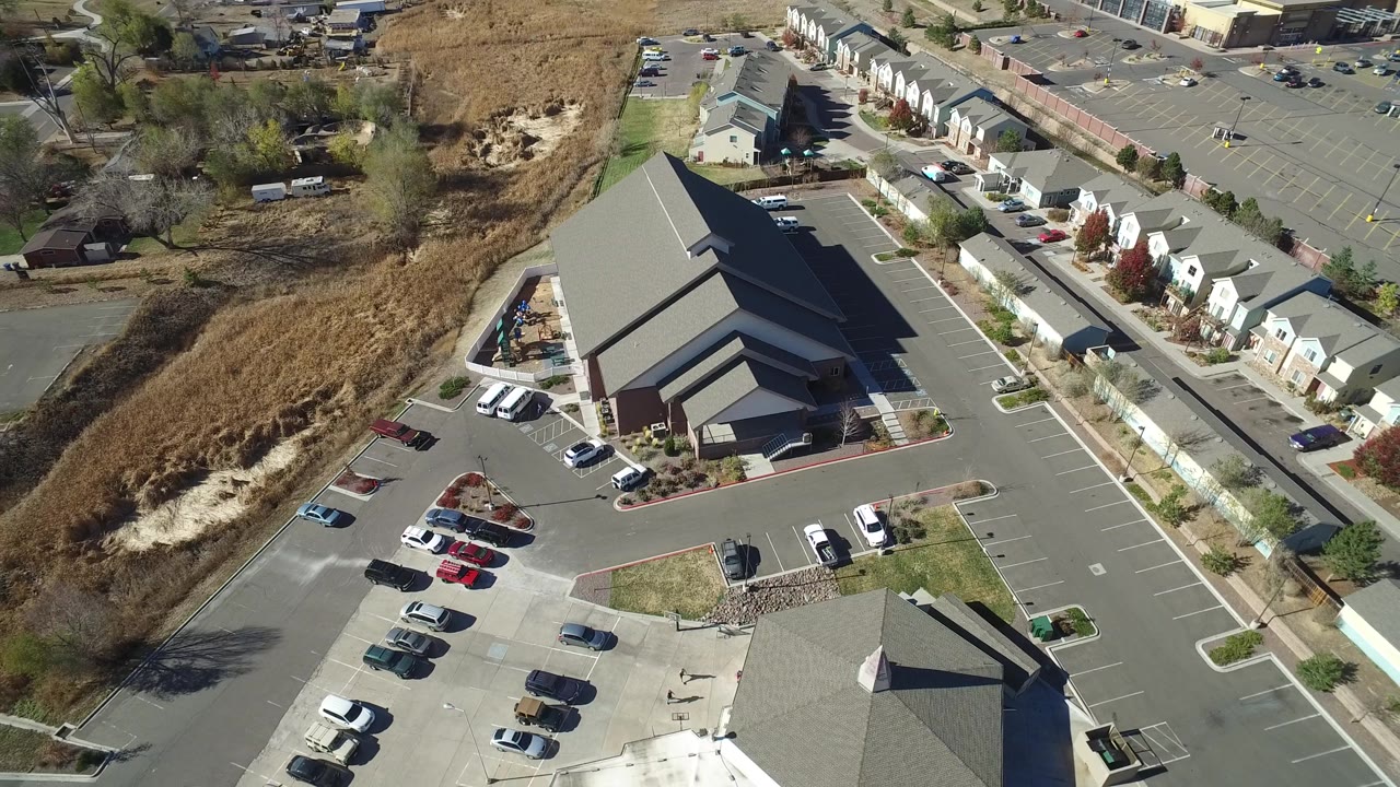 Drone-like overhead view of a chapel campus with parked cars, paved lanes, townhomes, and open land.
