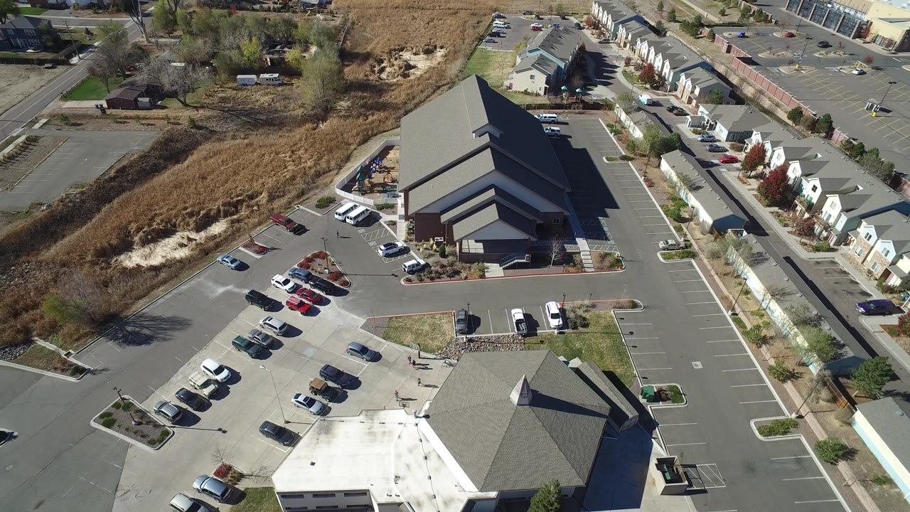 Aerial frame from Hidden Lake, Colorado showing a large angular dark-roofed building, parked cars, adjoining roads, dry open ground, nearby homes, and no visible people.