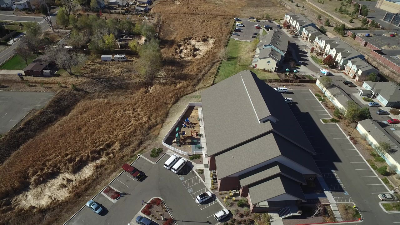Aerial view of a gray-roofed building, parking lot, townhomes, trees, and dry grassland.