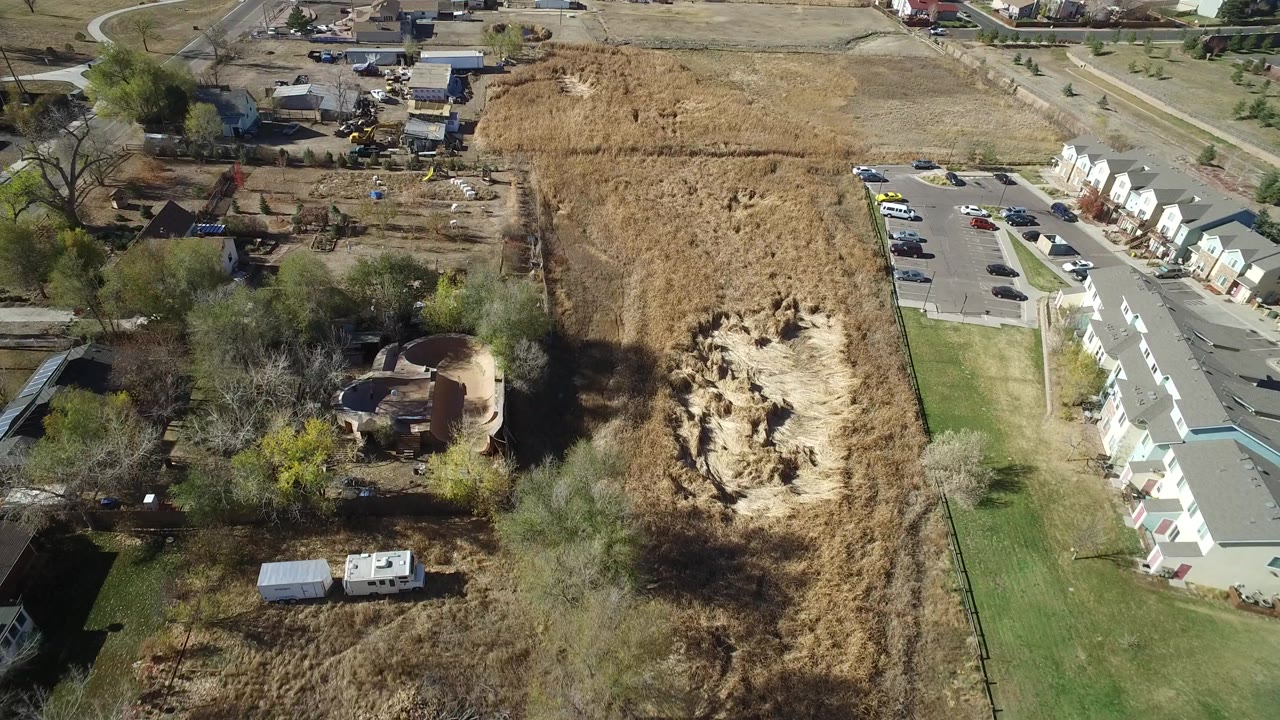 Second aerial frame in Hidden Lake, Colorado with the dirt pit centered between dry grass, trees, a residential row, parked cars, and no visible people.