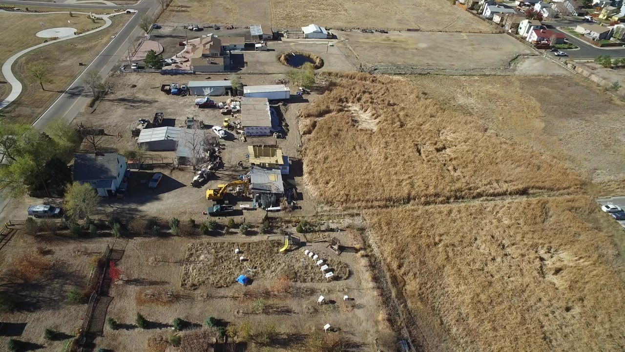 High aerial frame over Hidden Lake, Colorado showing a road, work sheds, parked vehicles, heavy equipment, dry grass lots, and no visible people.