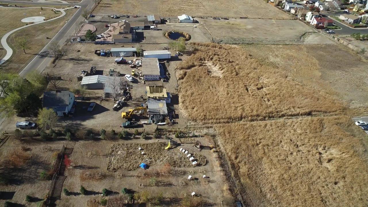 Aerial view in Hidden Lake, Colorado with utility buildings, a yellow machine, parked trucks, tan grassland, a paved road, and no visible people.