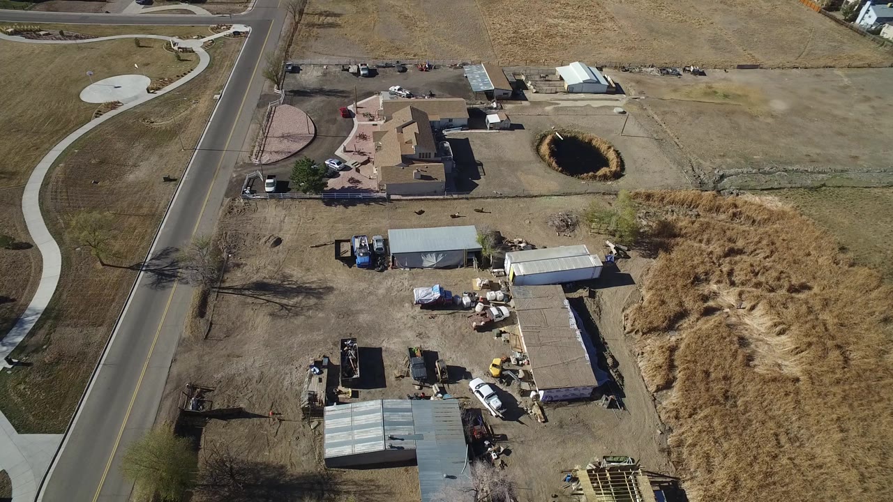 Aerial frame in Hidden Lake, Colorado with the road running vertically, a house cluster near the center, open dry field above, and a work yard with sheds below.