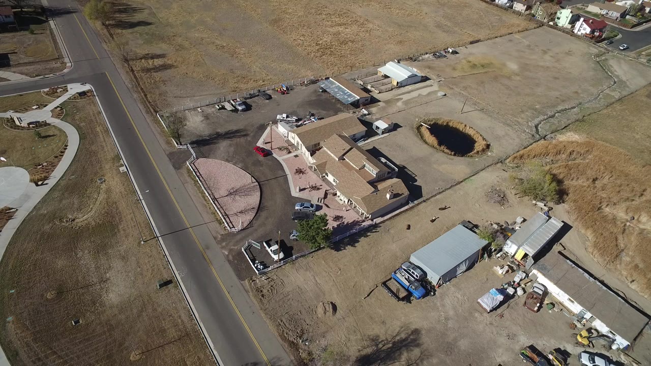 Aerial view of a road beside homes, dry lots, sheds, vehicles, and a small pond.