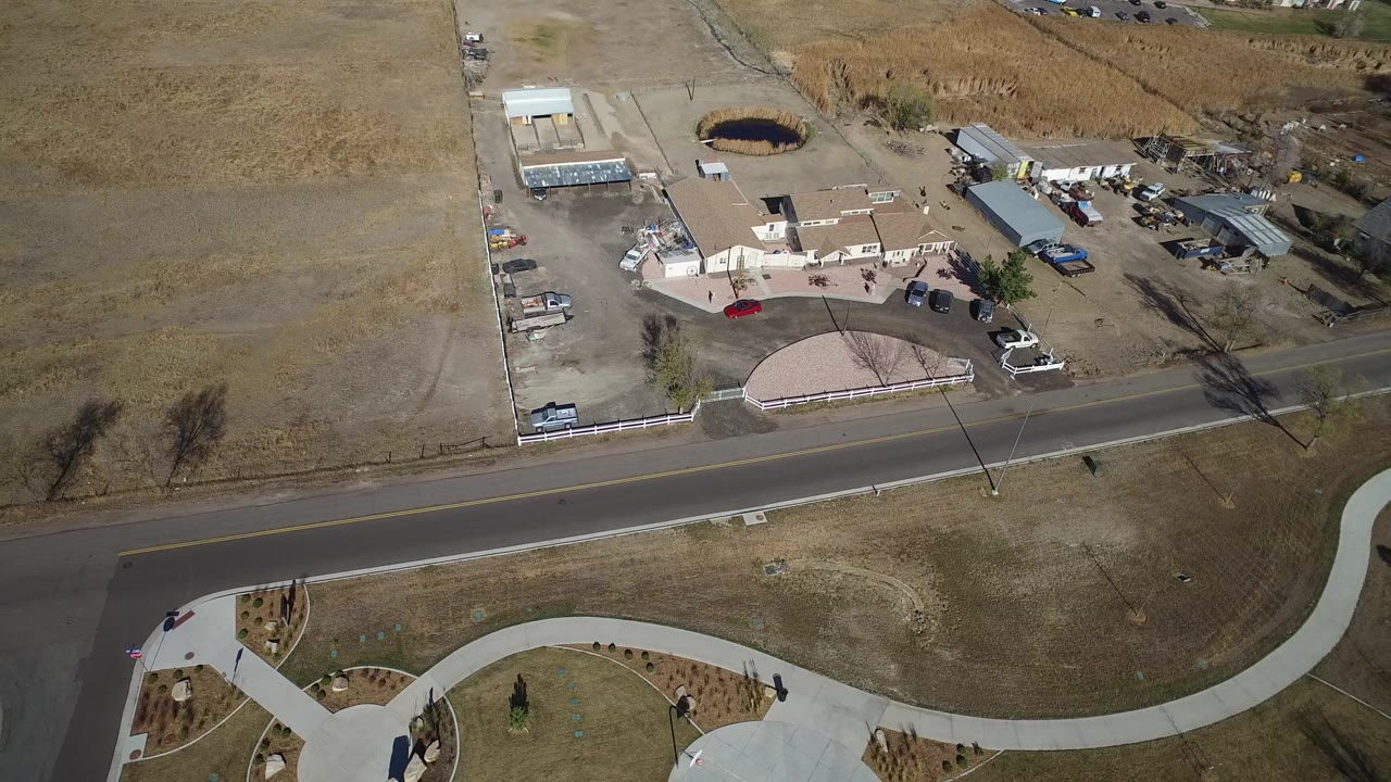 A high aerial frame over Hidden Lake, Colorado shows dry tan ground, a diagonal paved road, curving paths, a fenced property with low buildings, parked vehicles, and no visible people.