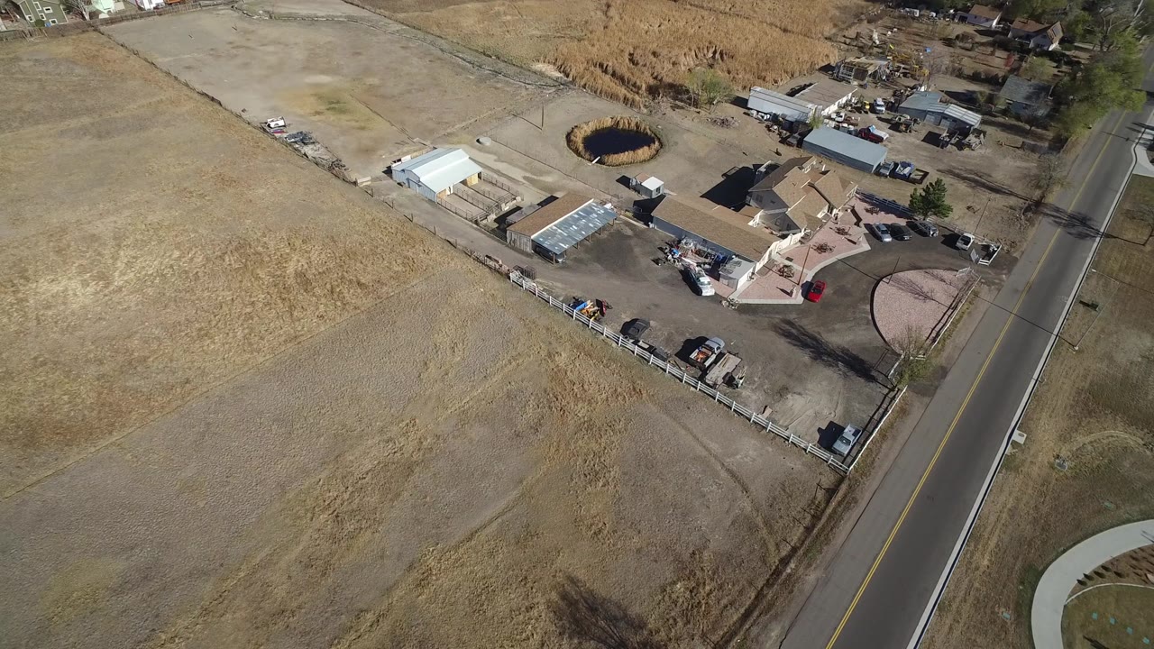 Aerial frame over Hidden Lake, Colorado with the road near the right edge, brown fields in the foreground, small buildings and parked vehicles beyond them, and no visible people.