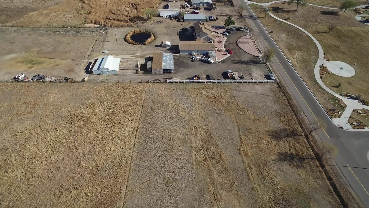Aerial view of dry fields, homes, sheds, road, and a dark circular pond or depression.