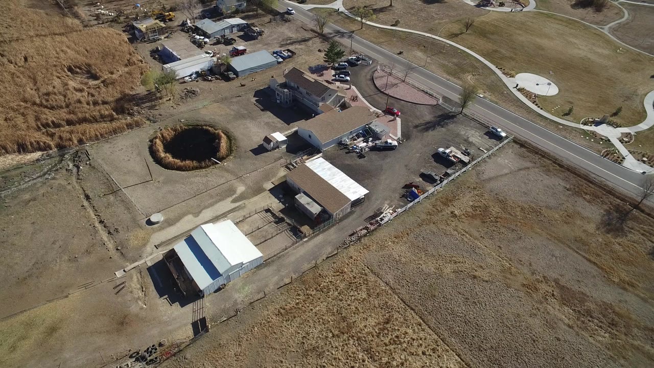 Aerial frame over Hidden Lake, Colorado with the curved road higher in frame, the pond left of center, white-roofed outbuildings below, and no visible people.