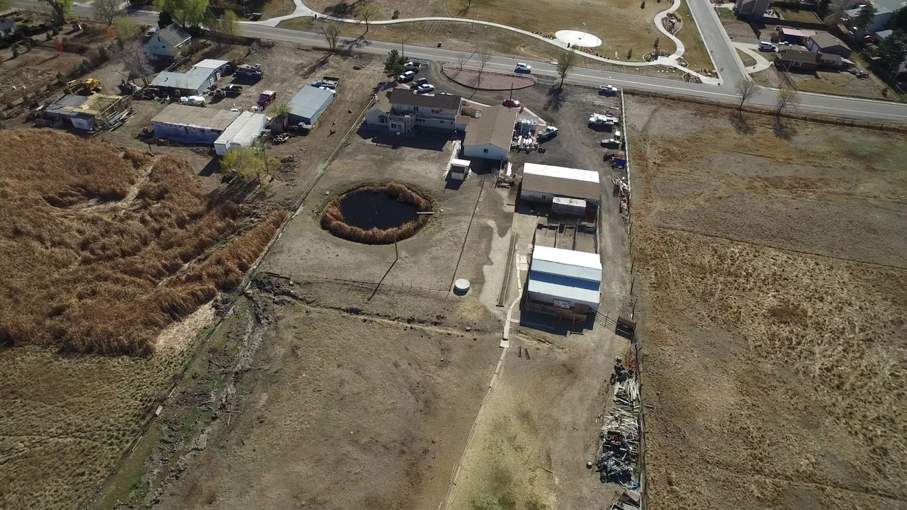 Aerial view in Hidden Lake, Colorado with the pond left of center, a pale dirt drive, sheds or outbuildings, roadside homes, and no visible people.