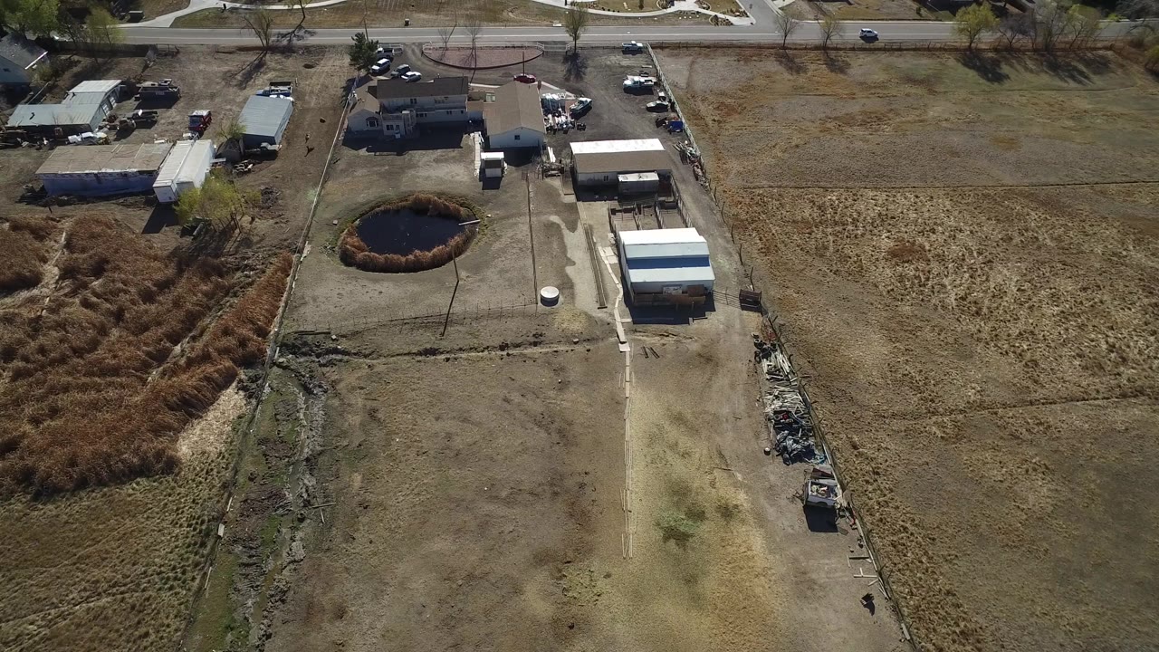 Aerial view in Hidden Lake, Colorado with the pond left of center, a long driveway, several sheds, private homes near the road, dry open field to the right, and tiny distant people near the road area.