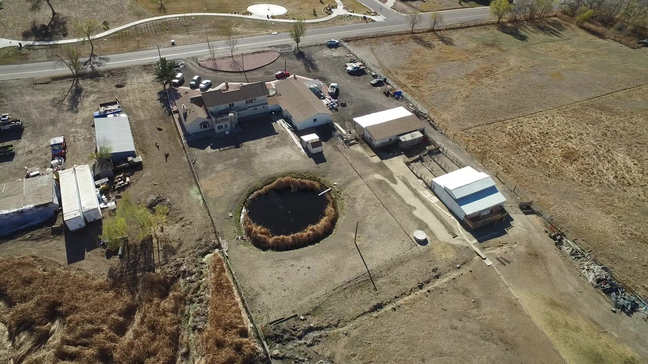 High aerial view in Hidden Lake, Colorado showing a tan-roofed rural inn property, outbuildings, parked vehicles, dry fields, a road, and a large dark round hole in the yard with no visible people.