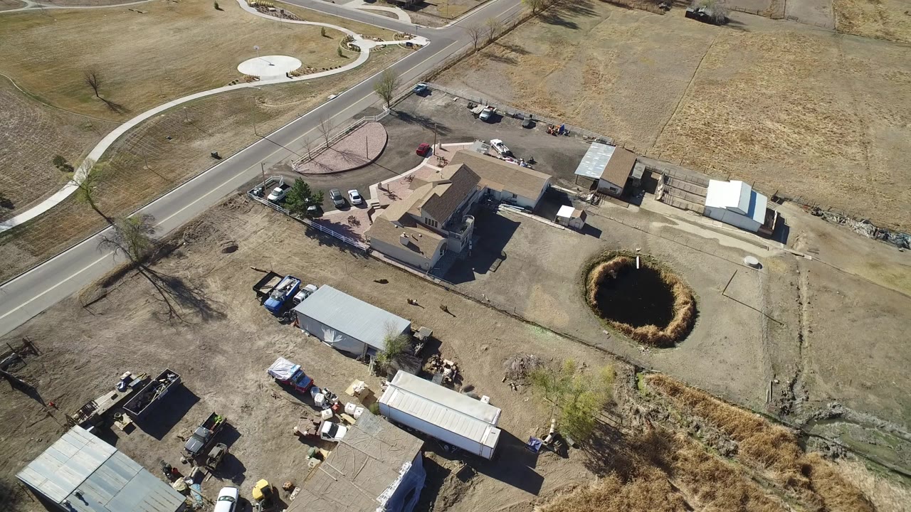 Overhead view in Hidden Lake, Colorado showing a tan house near a paved road, storage buildings, dry fields, and a small dark pond, with no visible people.