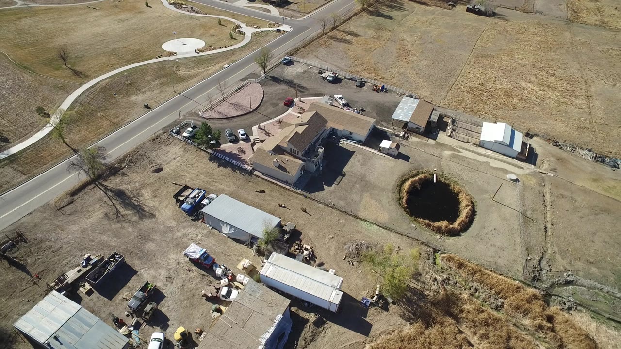 Aerial frame over Hidden Lake, Colorado with the house centered near a curved road, metal-roof sheds below, a pond to the right, and no visible people.