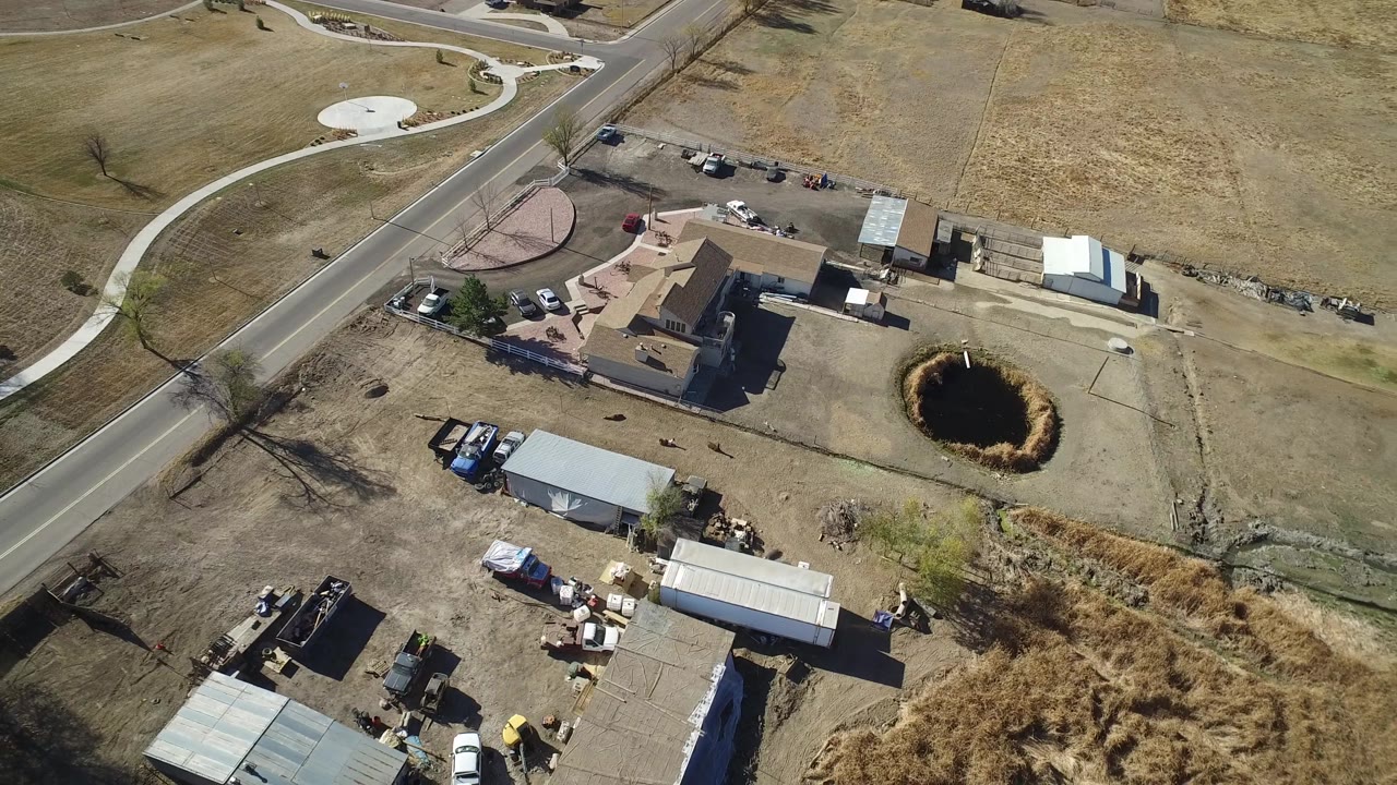 Drone-style view of a house lot, outbuildings, storage yard, pond, and surrounding fields.