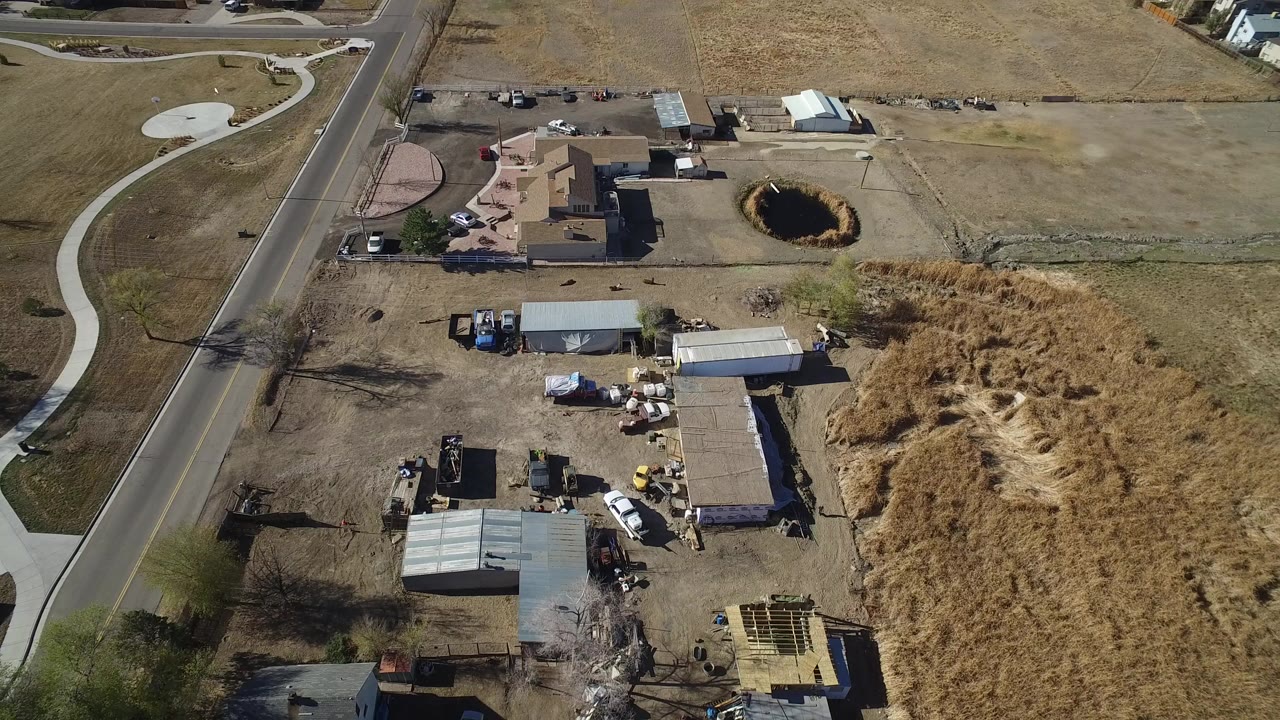 Opening aerial frame over a dry work site in Hidden Lake, Colorado, with sheds, parked vehicles, a nearby road, residential lots, tan grass, and no visible people.