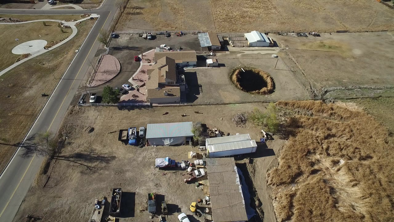 High overhead view in Hidden Lake, Colorado showing a paved road, tan open ground, private homes, metal sheds, parked vehicles, and a dark circular ground hole with no visible people.