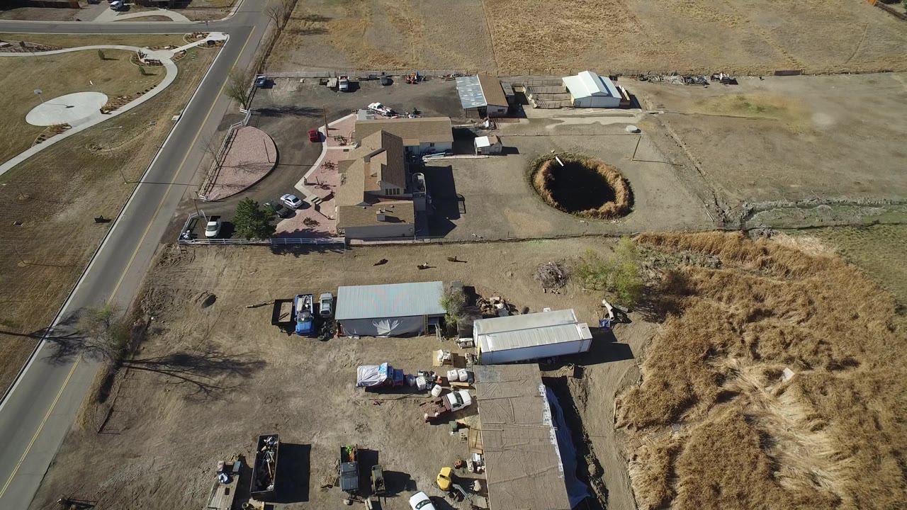 Drone-height frame over Hidden Lake, Colorado with the road running vertically, dry fields around the lots, outbuildings near parked vehicles, and the dark ground hole to the right with no visible people.
