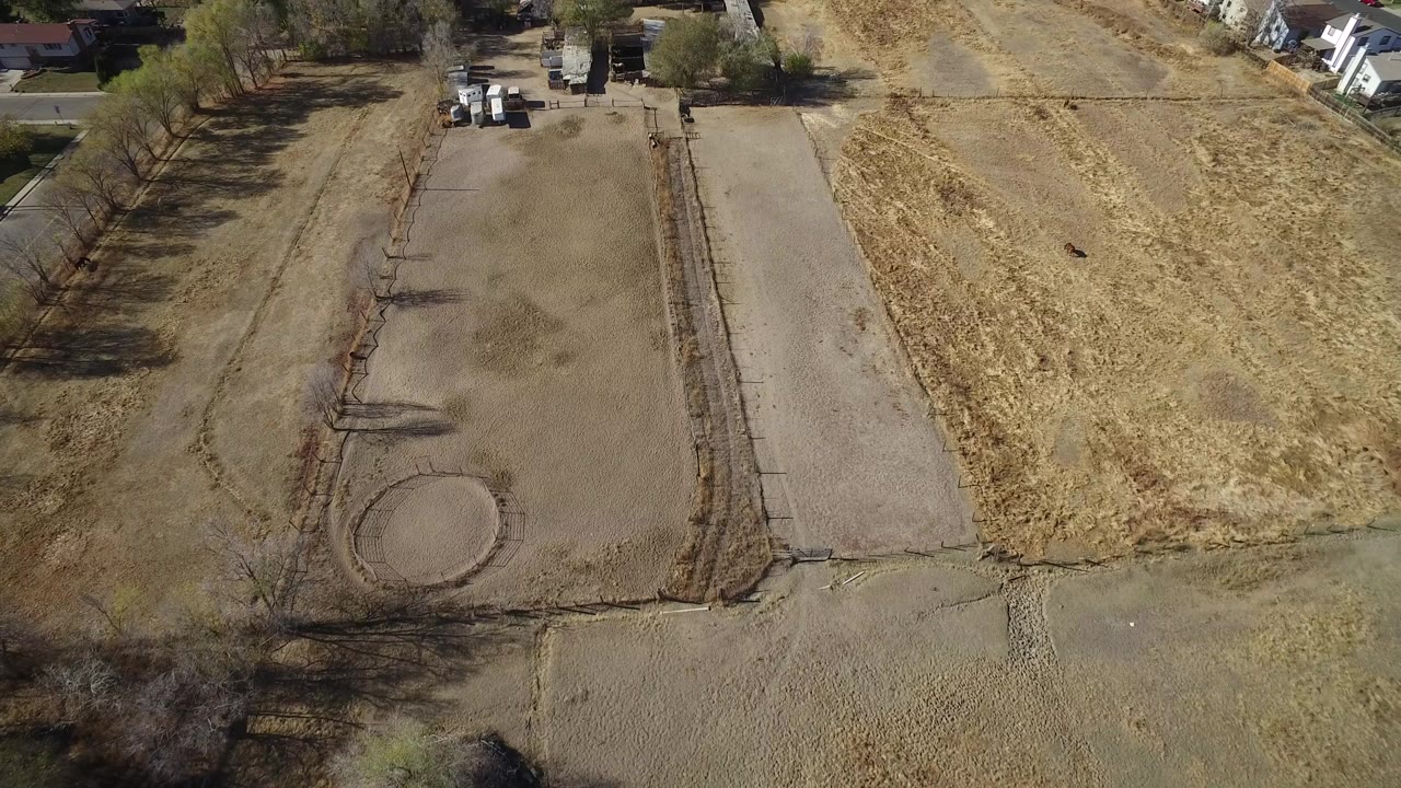 Opening overhead frame in Hidden Lake, Colorado showing dry rectangular field areas, a pale road, parked vehicles near trees, distant houses, and no visible people.