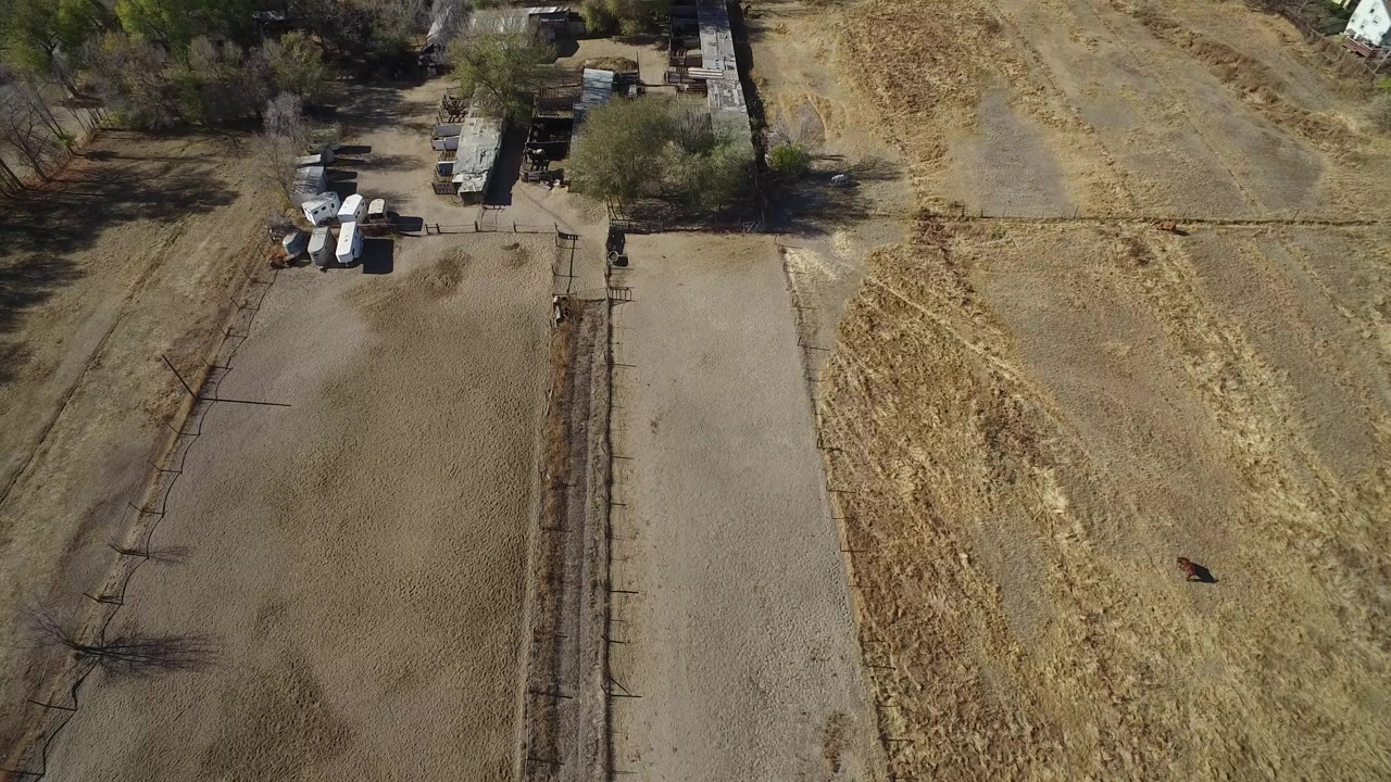 Aerial view of dry fields, a dirt lane, fences, trees, and small structures.