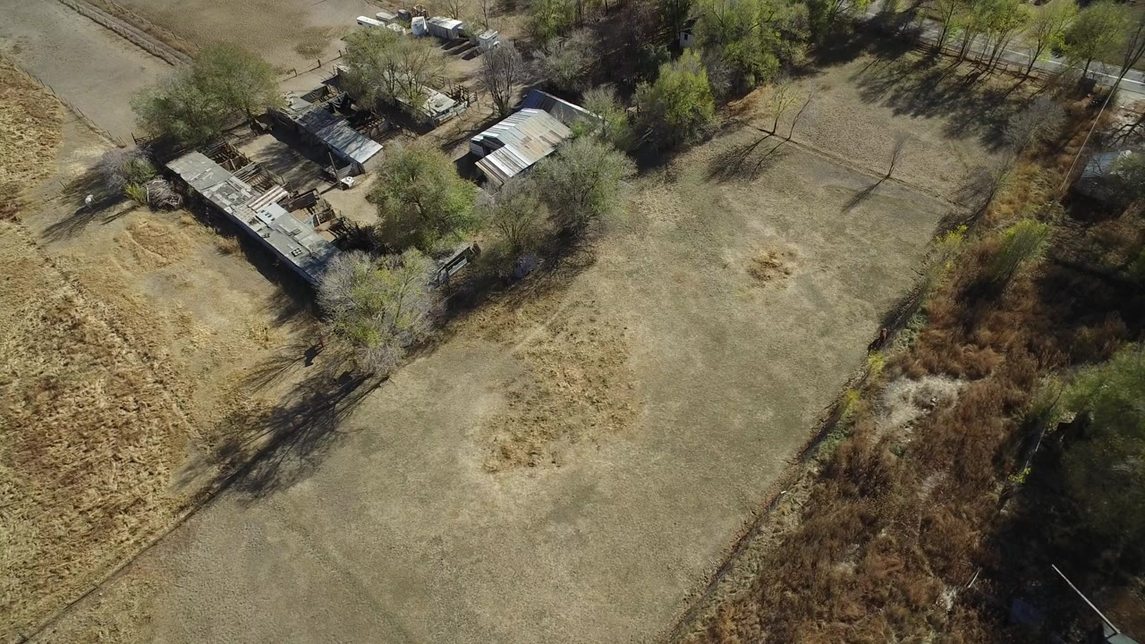 Opening aerial frame over a dry vacant lot in Hidden Lake, Colorado, with small outbuildings near the upper left, trees around the edges, and no visible people.