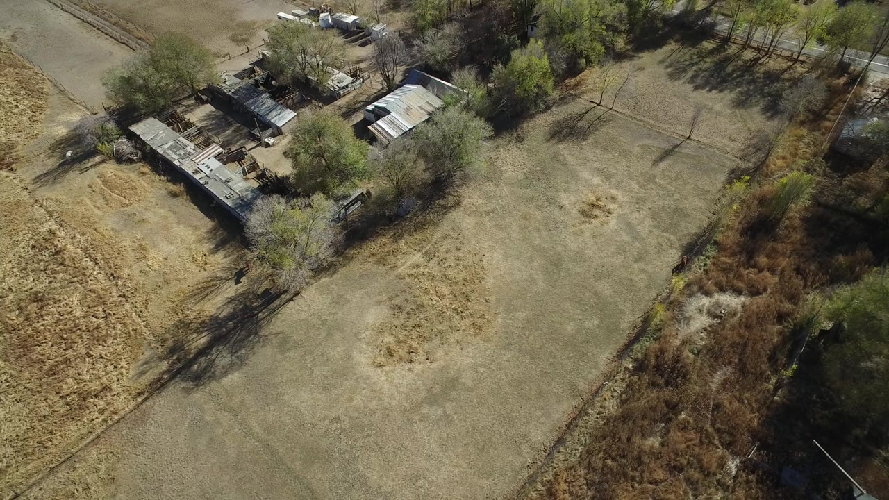 Aerial frame in Hidden Lake, Colorado showing open brown ground in the center, a brushy drainage or tree line along the lower right, small structures at the top, and no visible people.