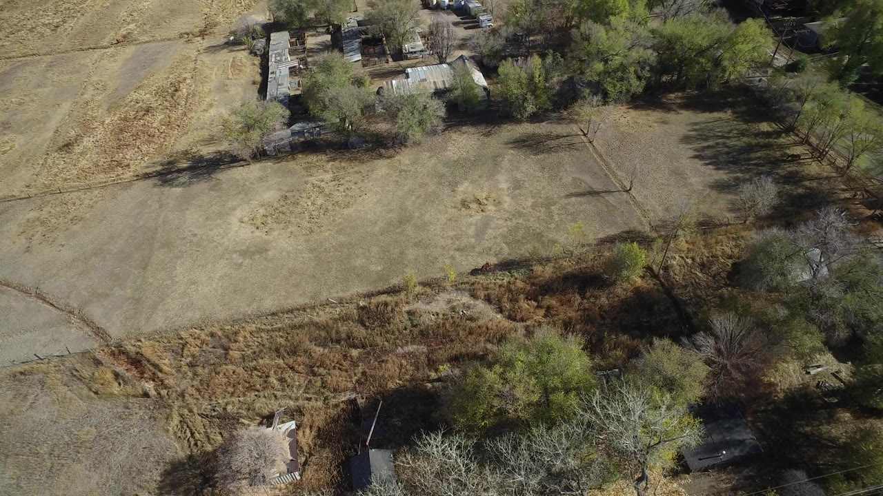 Opening aerial frame over Hidden Lake, Colorado, with a tan dry field centered below, tree shadows crossing the ground, small structures along the upper edge, and no visible people.