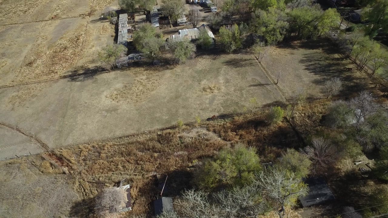 Second aerial frame over Hidden Lake, Colorado, showing the barren field slightly shifted in view, with brown brush in the lower area, trees along the right side, distant small buildings, and no visible people.