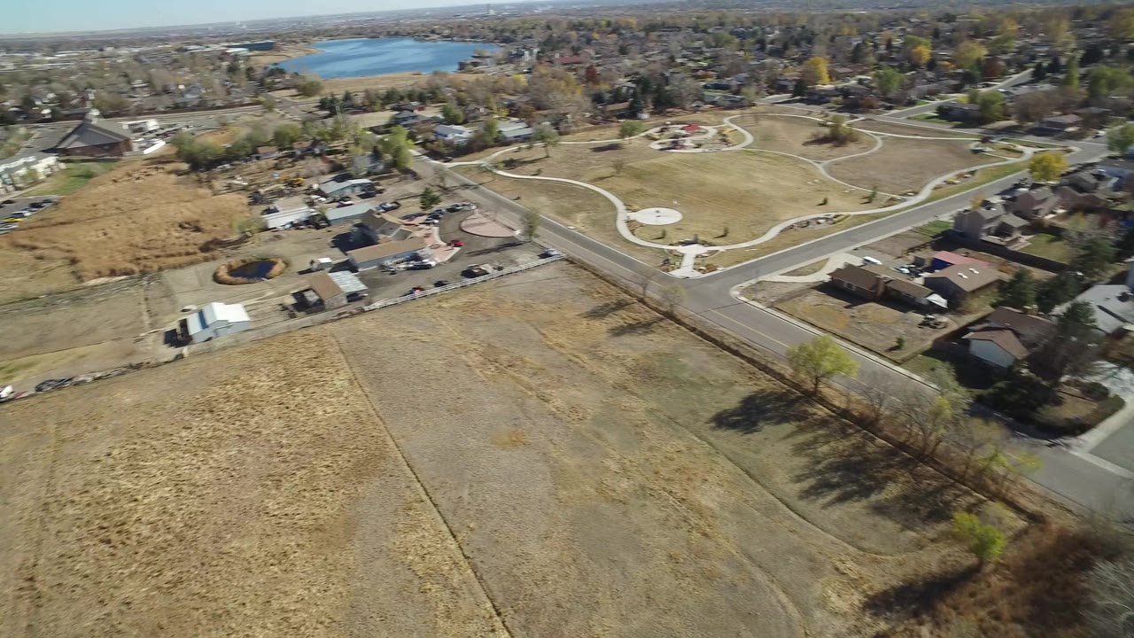 Hidden Lake, Colorado aerial frame with open tan fields closest to camera, a two-lane road and houses through the middle, curving park paths to the right, and distant water under a pale sky.