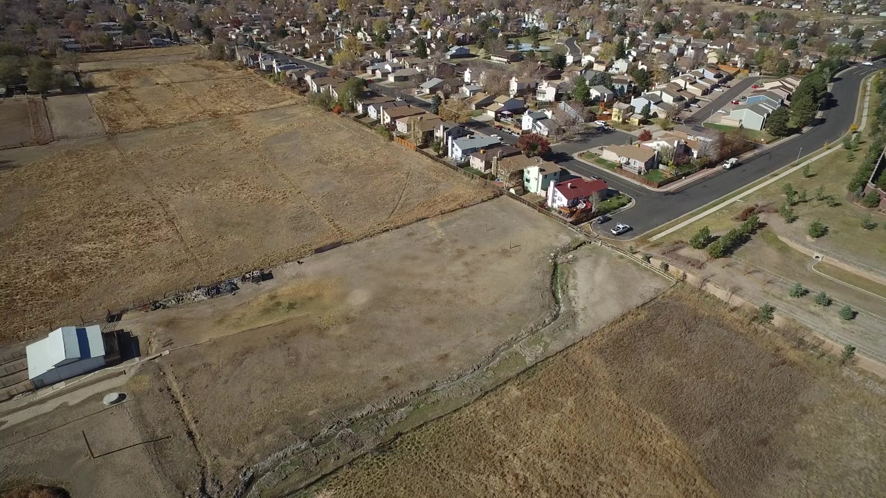 Aerial view in Hidden Lake, Colorado with tan fields across the lower frame, a road bend near the center, a small light-roofed structure at lower left, and no visible people.