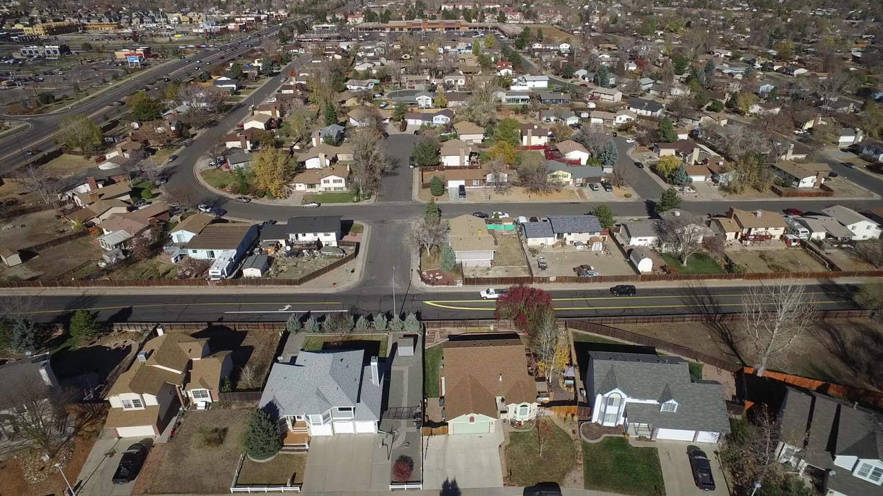 A high aerial view in Hidden Lake, Colorado frames rooftops and front yards in the foreground, a two-lane road across the center, and dense residential blocks beyond, with no visible people.